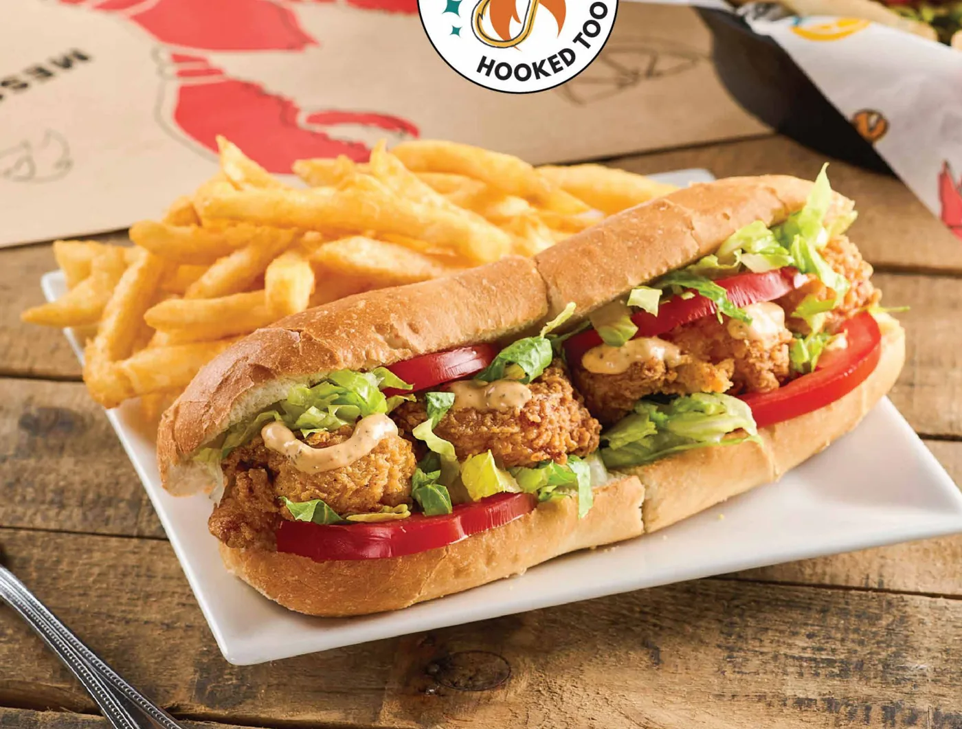A crispy fried po' boy sandwich with lettuce and tomato on a white plate, with a pile of fries